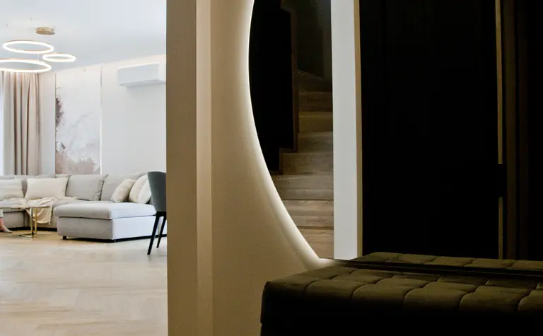 Nowoczesny korytarz z podświetlanym, okrągłym przejściem do salonu, w którym znajduje się jasna sofa i designerskie lampy wiszące. Schody i elegancka ławka w tle dodają wnętrzu stylu.