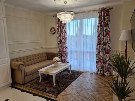 Tradycyjny salon z beżową sofą, kwiatowymi zasłonami i drewnianą podłogą.