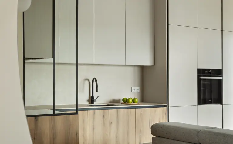 Nowoczesna kuchnia w stylu minimalistycznym z drewnianymi frontami, czarnym kranem i jasnymi szafkami, w tle wbudowany piekarnik. Przestrzeń w stonowanych barwach z eleganckim oświetleniem i akcentami ze szkła.
