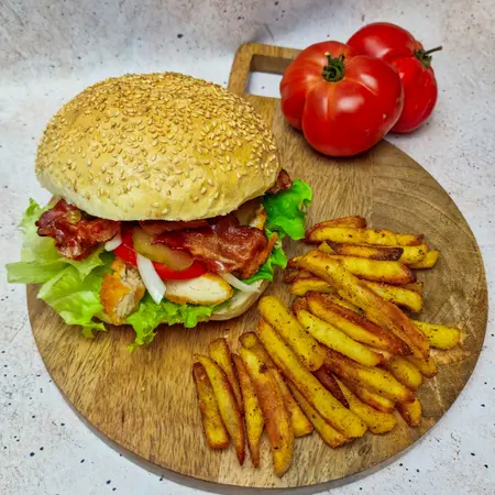 Burger z bekonem, sałatą i pomidorem na desce, obok frytki.