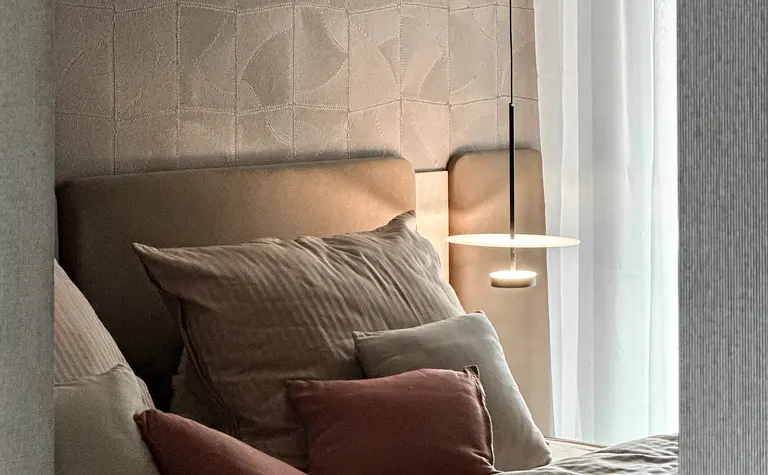 Wnętrze sypialni z beżowym łóżkiem, poduszkami i szarym tłem.