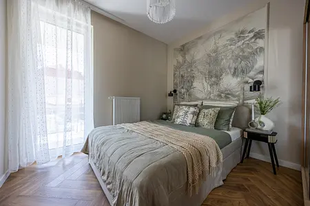 Przytulna sypialnia z dużym łóżkiem w neutralnych odcieniach, ozdobiona tropikalną tapetą nad zagłówkiem, naturalnymi dodatkami i tekstyliami.