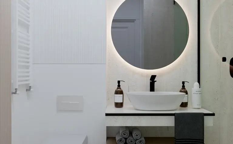 Wnętrze łazienki z okrągłym lustrem, minimalistycznymi meblami i detalami w stylu art deco i współczesnym.