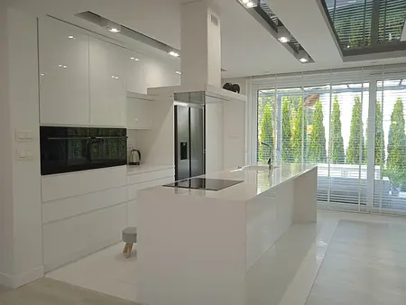 Jasna, nowoczesna kuchnia z dużą wyspą, białymi frontami i dużym oknem z widokiem na ogród. Minimalistyczne wykończenie i dużo naturalnego światła.